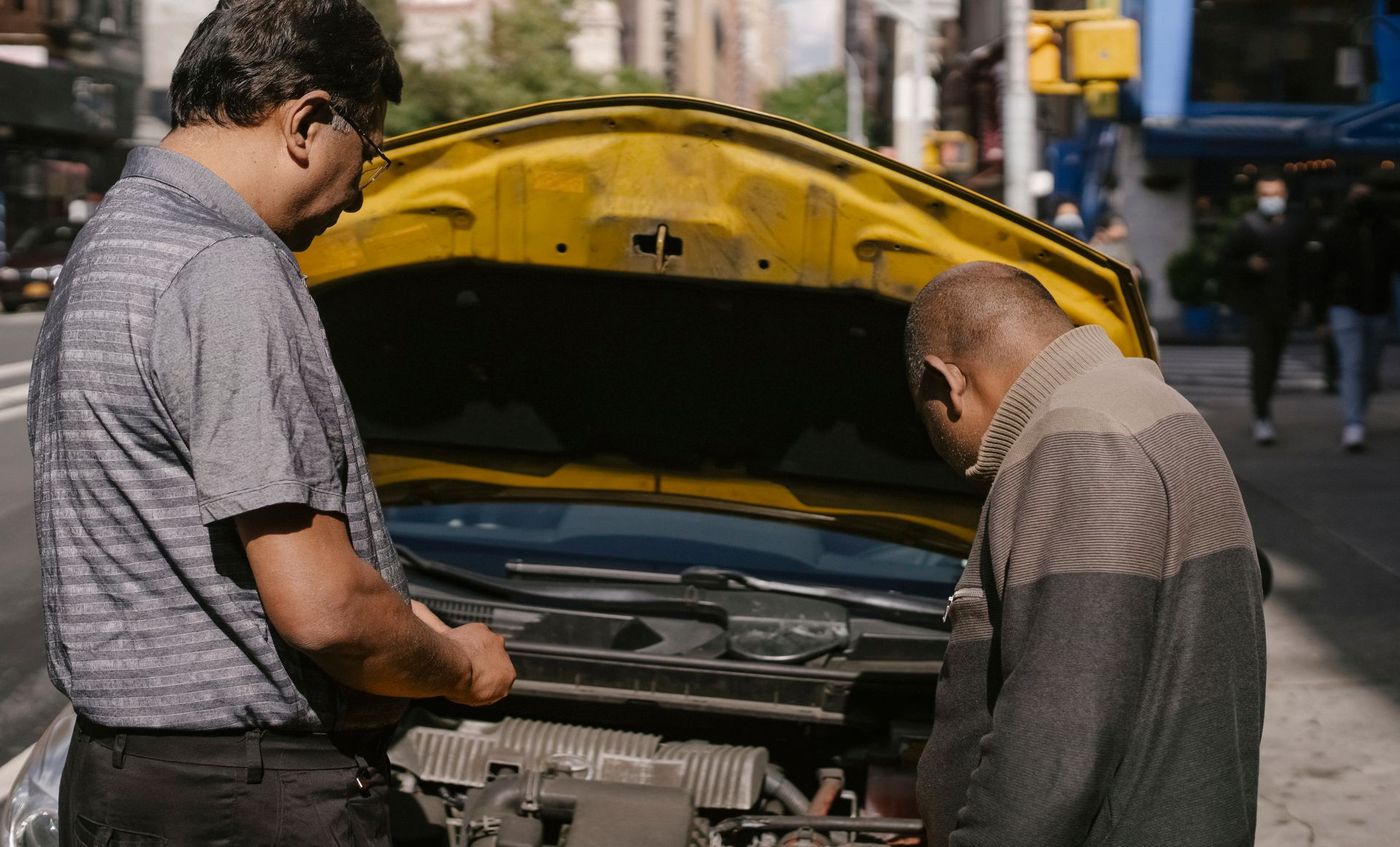 Two people looking at a car engine with the hood up at the side of a street